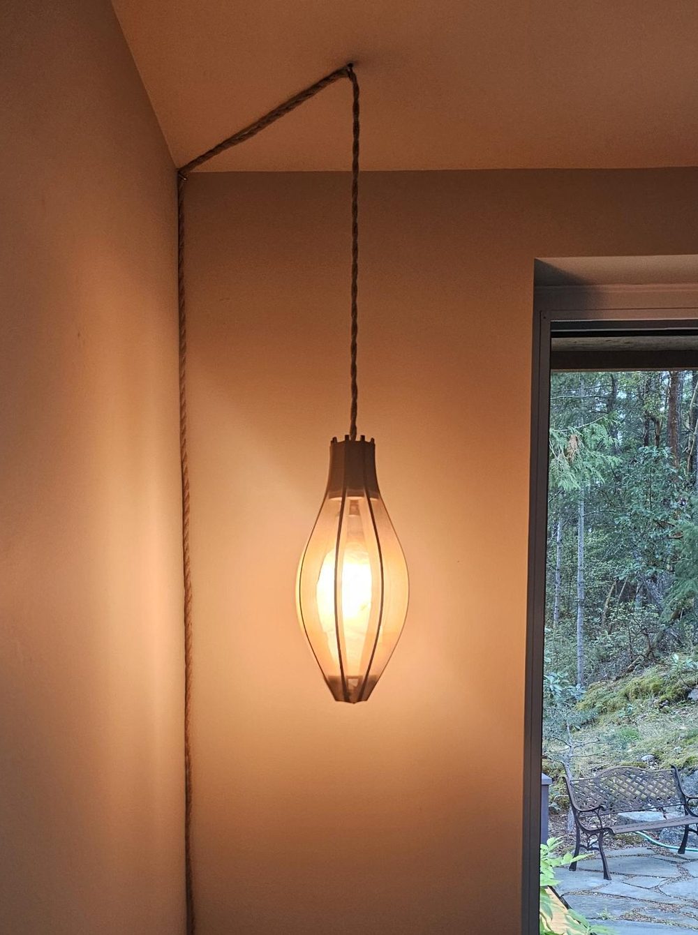 A ribbed oval wooden lamp hanging lit on a rope cord beside a window