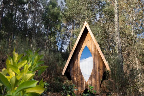 A small A-frame of charred timber with an almond-shaped window, on a wooded hillside
