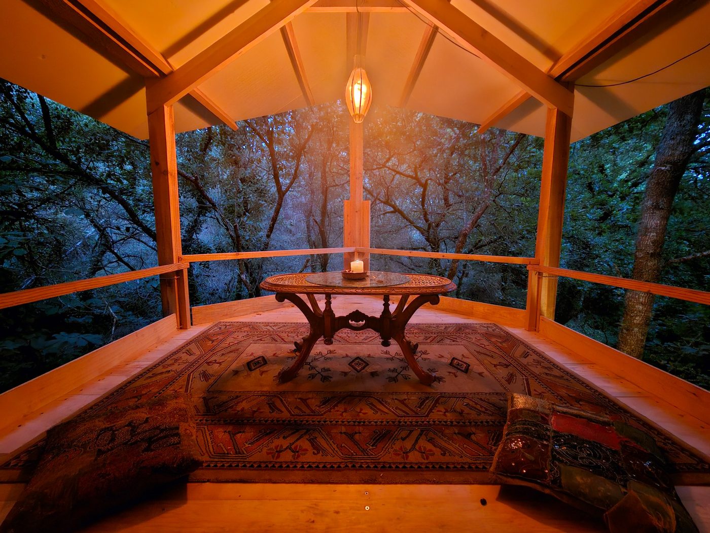 An open timber room among the trees at dusk, with a carved table, a rug and floor cushions under a vaulted ceiling lit by a wooden lantern